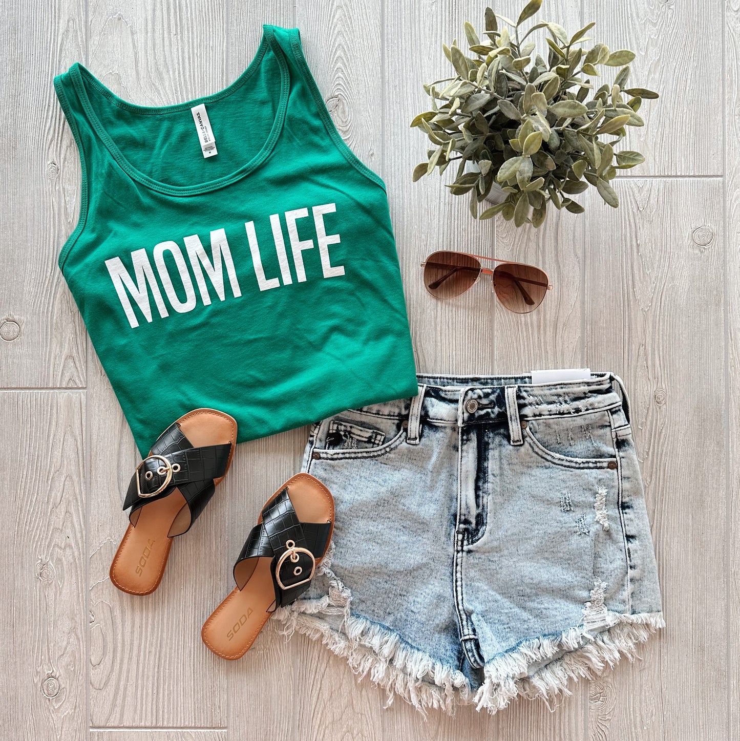 The MOM LIFE • Unisex Tank Top is styled with light denim cut-off shorts, black sandals, brown sunglasses, and a small potted plant on a light wooden floor.