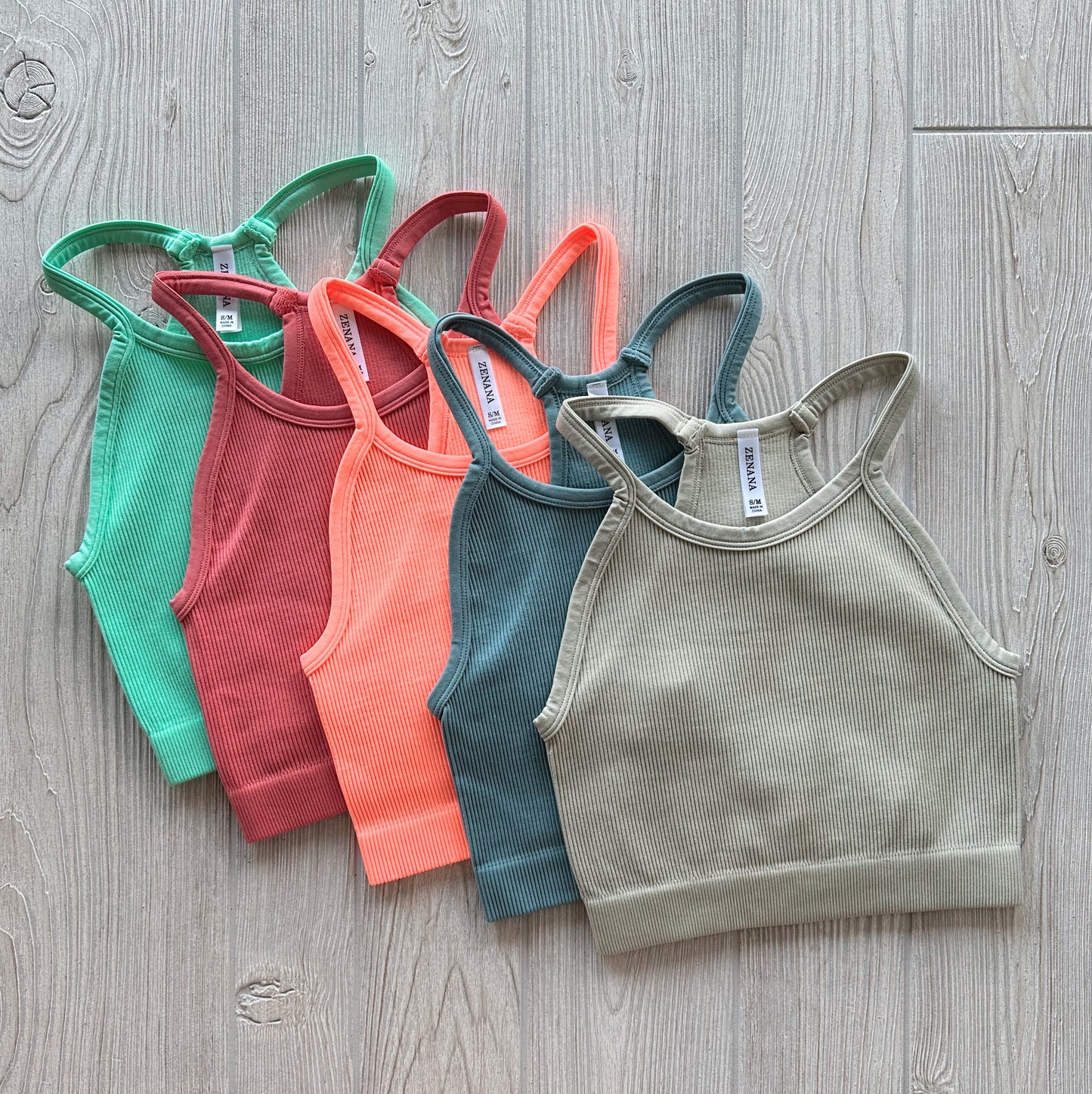 Five Luna Ribbed Crop tops from the Austin Collection in green, red, orange, blue, and beige are fanned out and overlapping on a light wood floor. Each features a ribbed racerback design with a high neck and stretchy fabric.