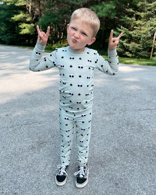 A young child stands outdoors in the Rad Skelly • 2 Piece Bamboo Set (Classic Fit) featuring matching skull and star print pajamas. The child makes a playful face, flashing a rock on gesture. Trees and greenery fill the background.