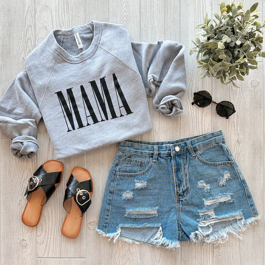 A flat lay of a casual outfit featuring the MAMA • Gray Bella Pullover, distressed denim shorts, black slide sandals, black sunglasses, and a small potted plant on a light wood floor.