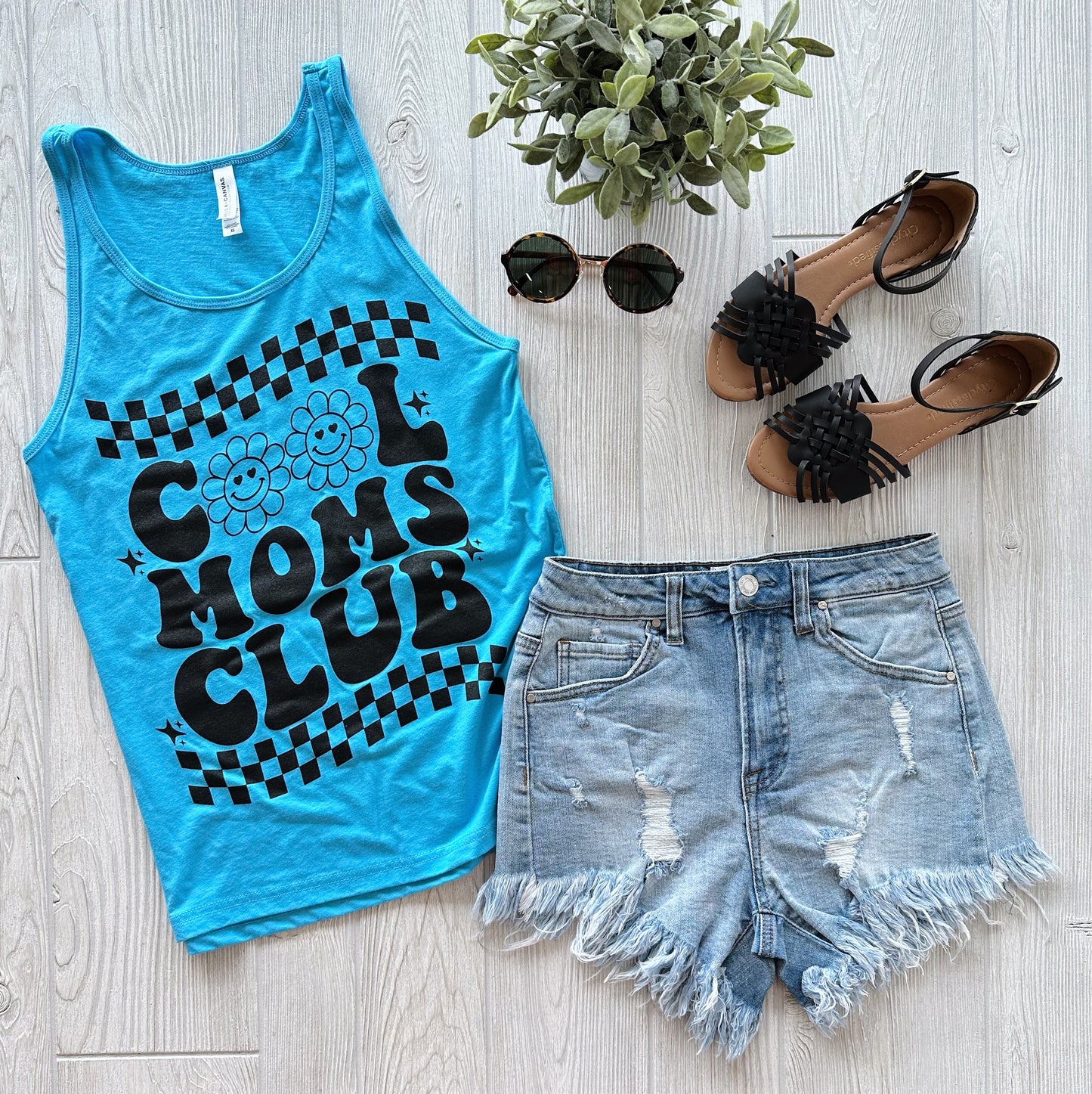 A flat lay of the Cool Moms Club • Unisex Tank Top in neon blue, styled with distressed denim shorts, black strappy sandals, round sunglasses, and a small green potted plant on a light wooden floor.