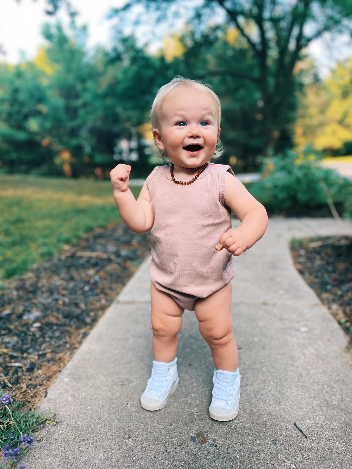 A smiling blond baby stands on a sidewalk outdoors, wearing the Organic Cotton Bubble Romper in sleeveless pink, paired with white sneakers and a beaded necklace. Green trees and grass are visible in the background.