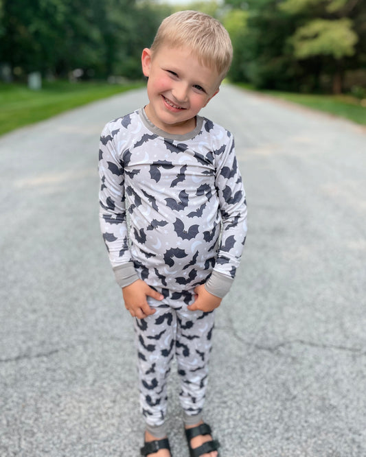 A young boy smiles on a paved road wearing the Batty • 2 Piece Bamboo Set (Classic Fit) in gray camouflage and black sandals, from our cozy collection. Trees and greenery fill the background.