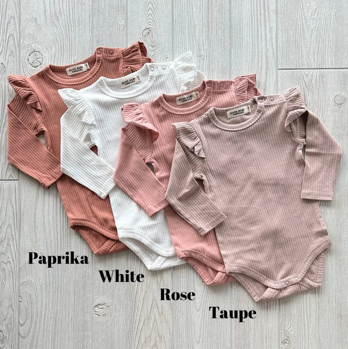 Four Ribbed Ruffle Bodysuits in paprika, white, rose, and taupe—made from organic cotton—are shown side by side on a light wood floor, each labeled by color. A perfect cool weather basic for your little one.