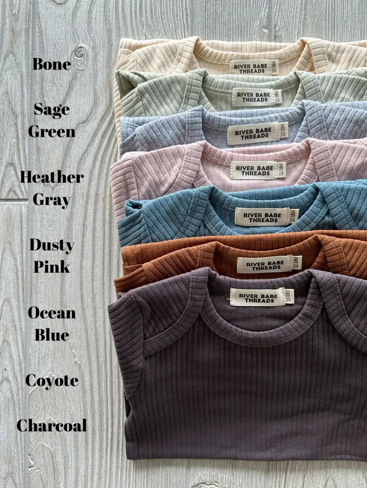 Seven Ribbed Long Sleeve Bodysuits in organic cotton—bone, sage green, heather gray, dusty pink, ocean blue, coyote, and charcoal—are neatly stacked on a light wood surface with their color names listed alongside.