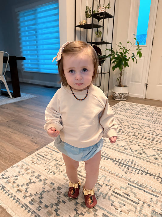 A toddler with light brown hair stands indoors on a patterned rug, wearing a beige French Terry Pullover, blue shorts, red shoes, and a beaded necklace. Behind them are plants, shelves, and a large window—a cozy cool weather look.