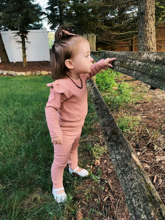 A young child stands outside by a wooden fence, pointing ahead, wearing Ribbed Organic Cotton Leggings with white shoes; grass and trees are in the background.