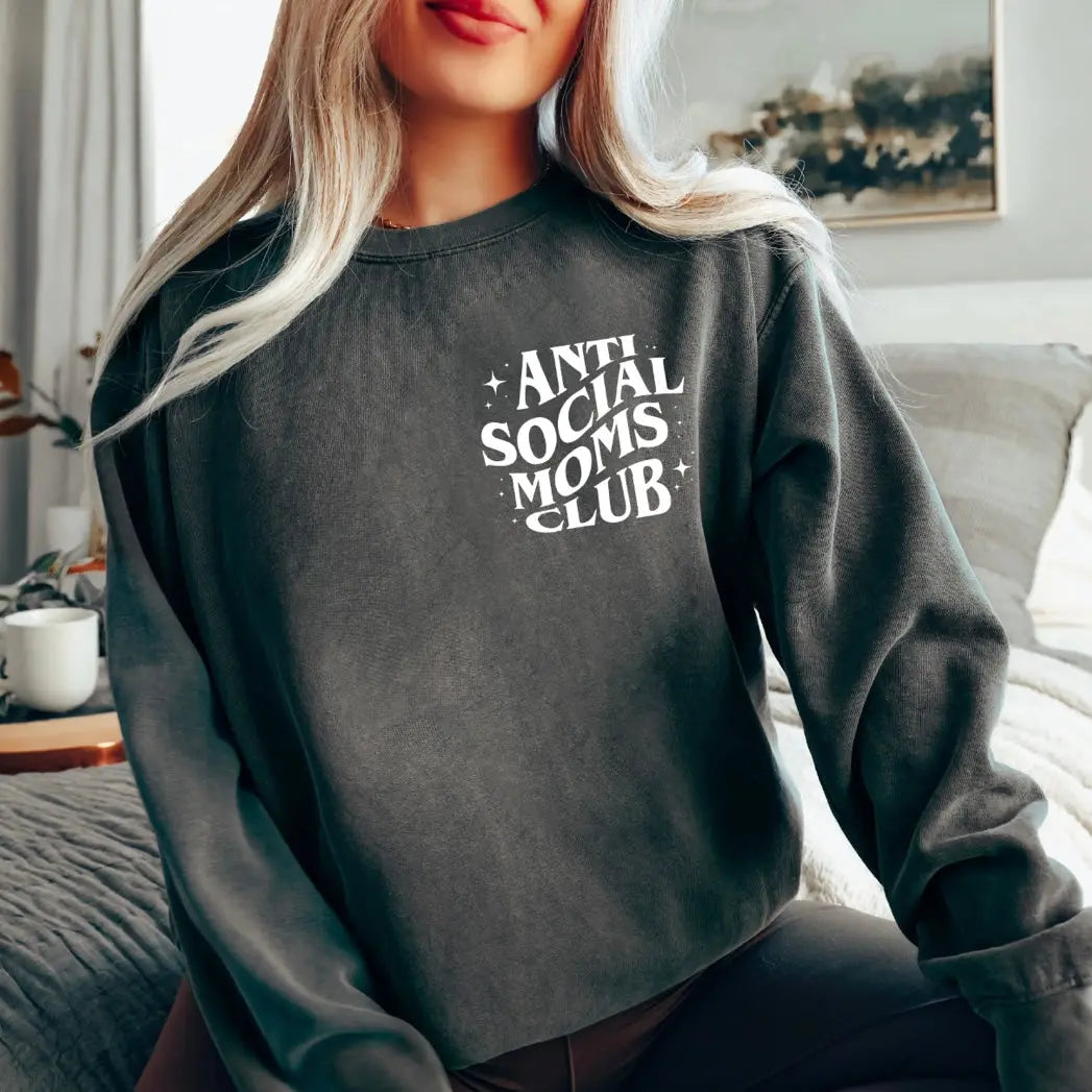 A woman with long blonde hair sits on a bed wearing the Anti Social Moms • Pepper Long Sleeve tee, featuring white Anti Social Moms Club text. The background shows bedding, a plant, and a framed picture on the wall.
