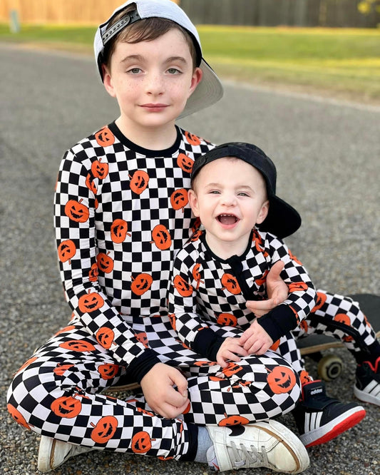 Two children wear matching Checkered Pumpkins • Bamboo Zippy (Classic Fit) outfits with orange jack-o’-lanterns and backward caps, smiling by a skateboard. The grow-with-me design ensures lasting comfort and style.