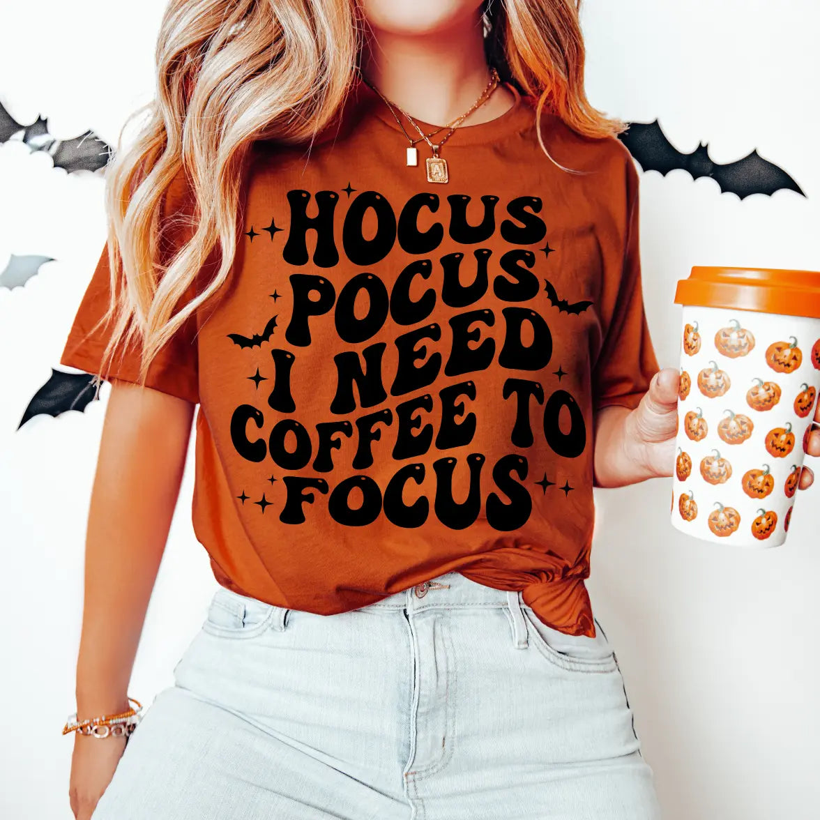 A woman wears the Coffee to Focus • Tee, featuring Hocus Pocus I Need Coffee to Focus, and holds a matching Hocus Pocus coffee cup. Black bat decorations create a spooky vibe on the white wall behind her.