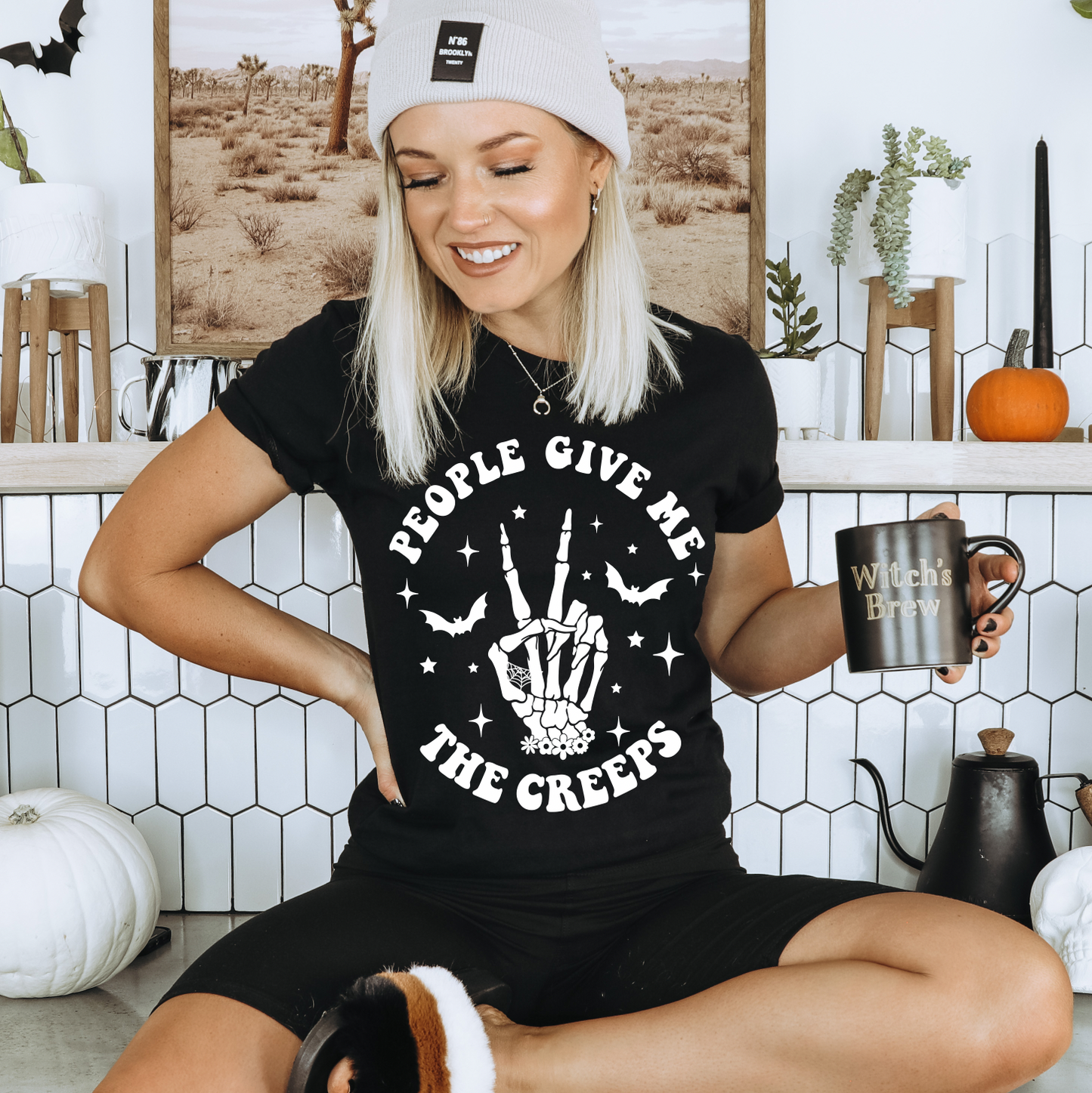 A smiling woman in a beanie and The Creeps • Black Tee, made from soft Airlume cotton, sits in a Halloween-themed kitchen. She holds a Witch’s Brew mug and is surrounded by white pumpkins.