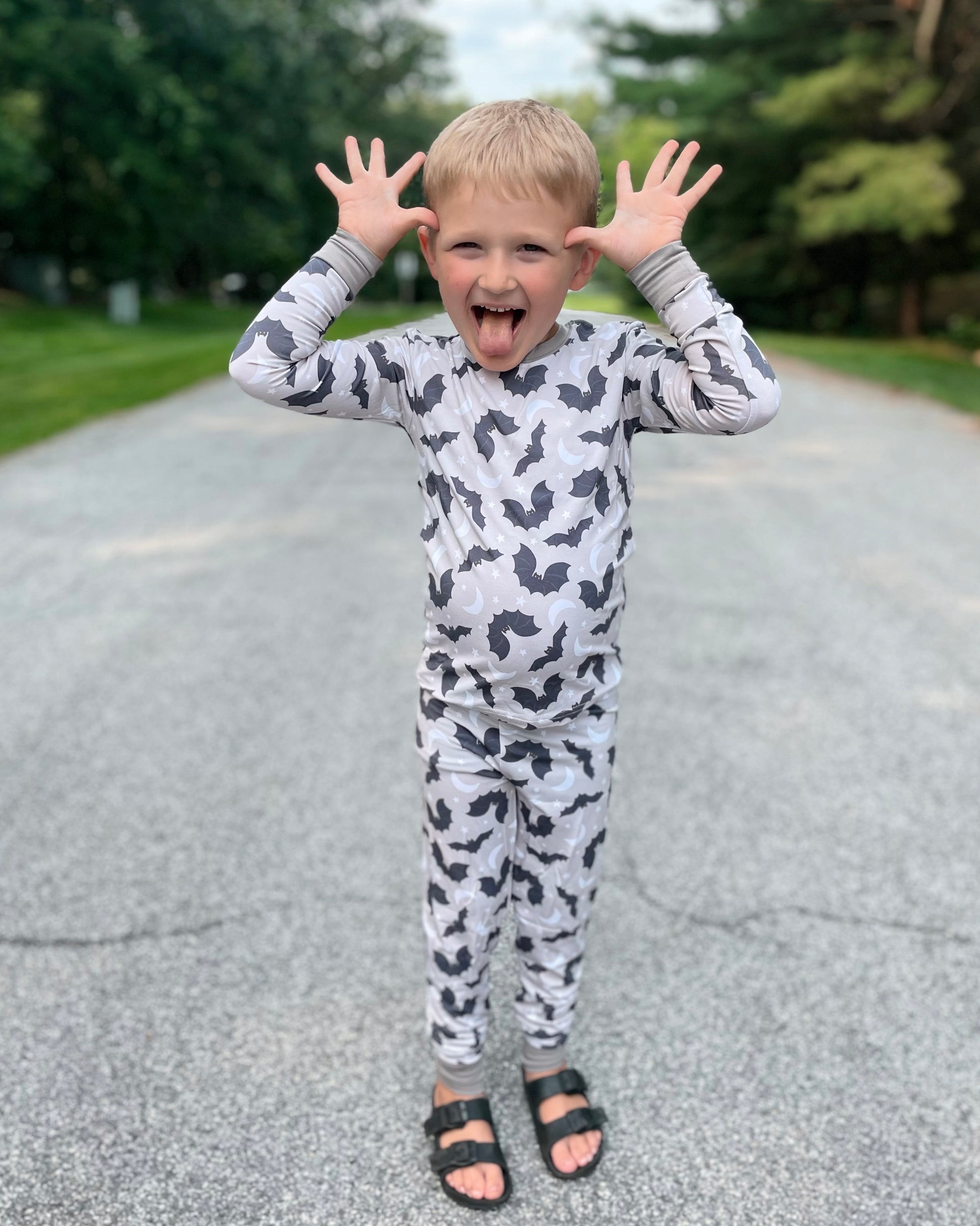 A young boy wears our Batty • 2 Piece Bamboo Set (Classic Fit) and stands on a paved road, making a silly face with his tongue out and hands raised like antlers. Trees and grass line the background.