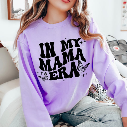 A person sits on a bed wearing the Mama Era • Orchid Long Sleeve, a pastel purple shirt with bold black text IN MY MAMA ERA and butterfly accents. The persons face is partly out of frame.