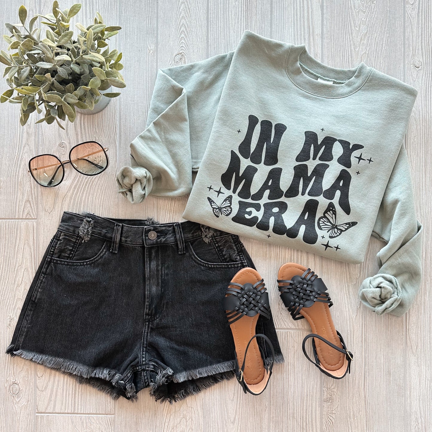 Flat lay of the MAMA ERA • Sage Pullover, black denim shorts, woven black sandals, round sunglasses, and a small potted plant arranged on a light wooden surface.