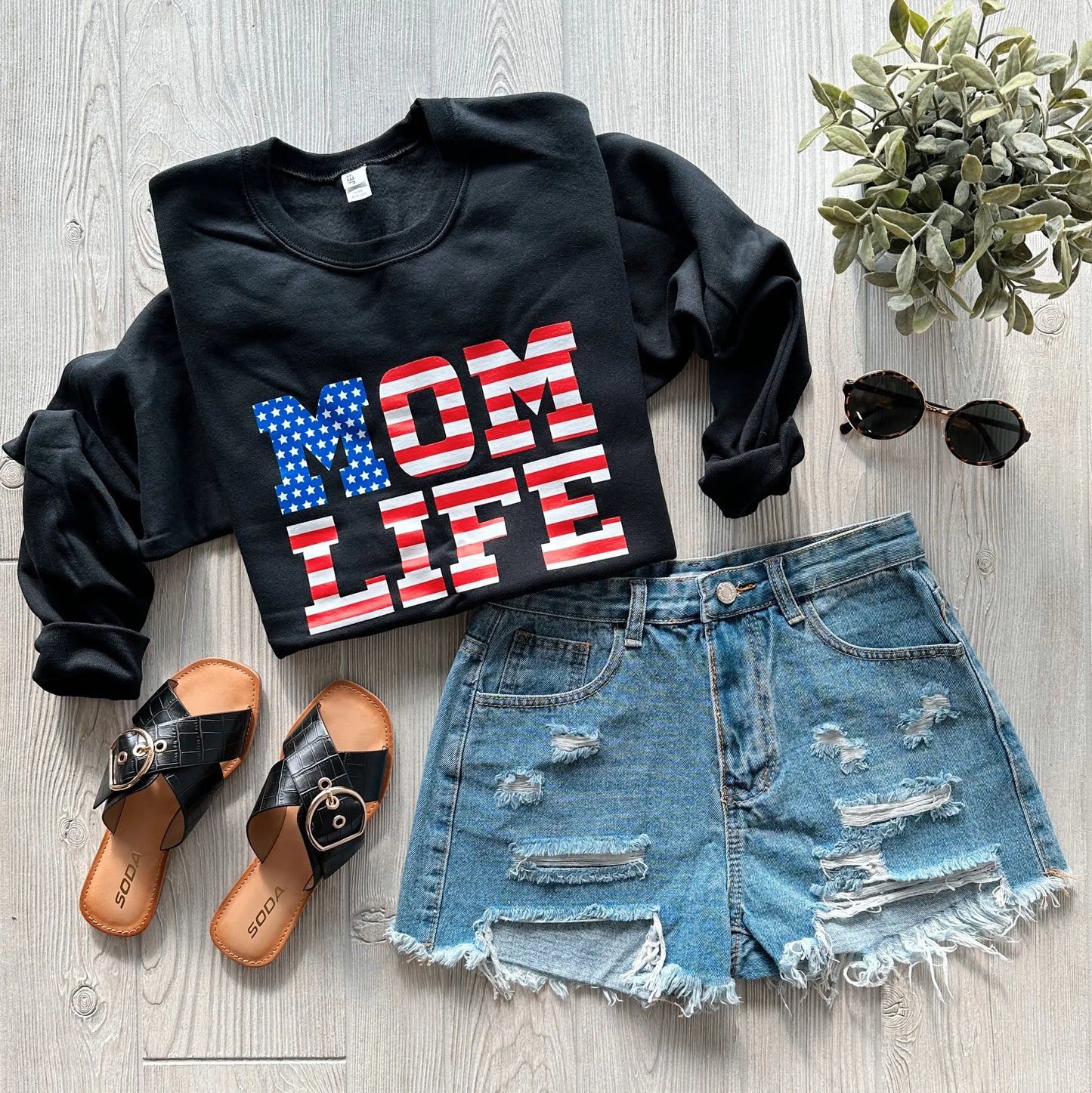 Flat lay of the Mom Flag Print • Pullover featuring “MOM LIFE” in an American flag design, styled with distressed denim shorts, black slide sandals, sunglasses, and a small potted plant on a light wood floor.