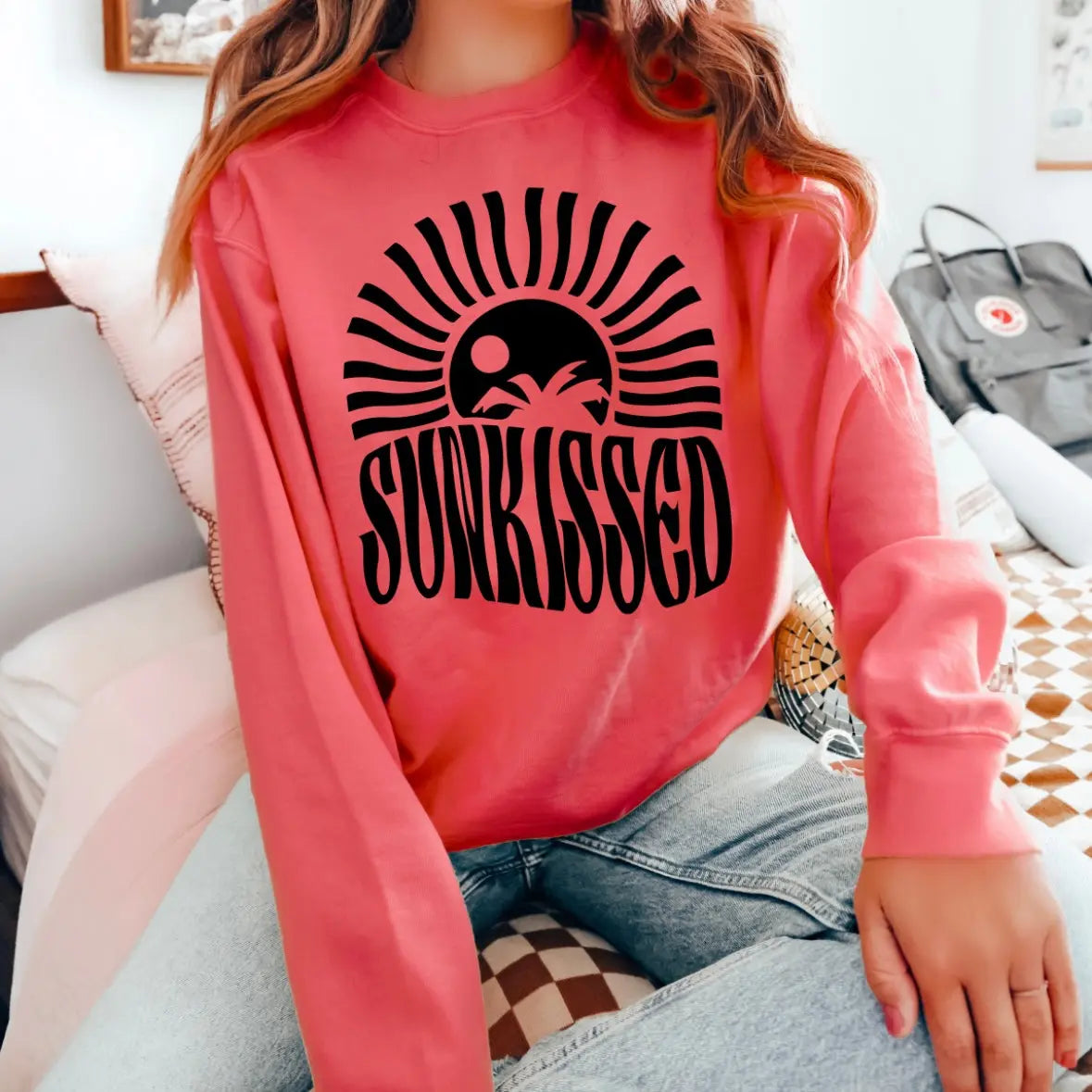 A person in light blue jeans sits on a bed, wearing the Sunkissed • Watermelon Long Sleeve tee featuring a black sun and palm trees graphic. A gray bag and books are on the bed in the background.