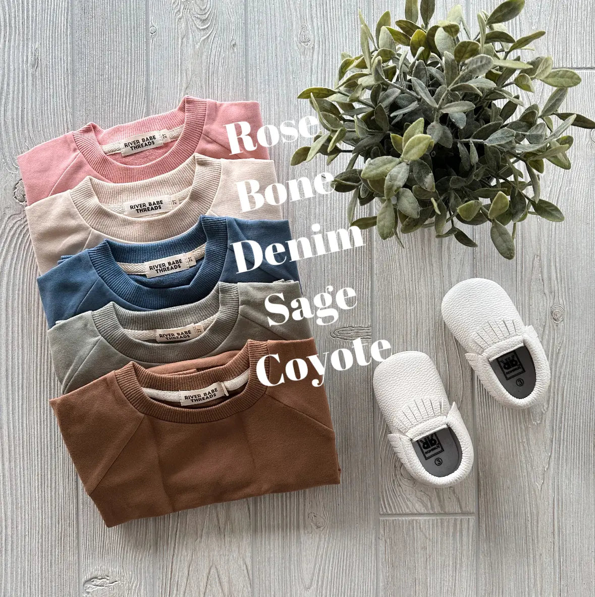 Five folded French Terry Pullovers in rose, bone, denim, sage, and coyote are stacked near white baby shoes and a potted plant on a light wood floor. Each color is labeled. Made from organic cotton—perfect for cool weather.