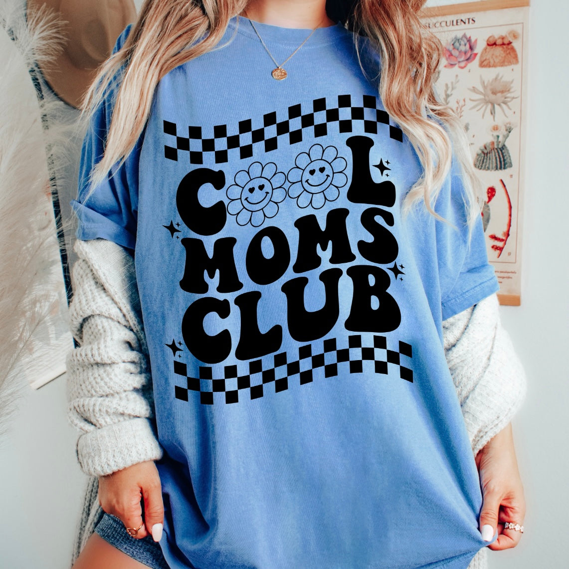 Woman wears the Cool Moms Club • Royal Dyed Tee—soft ringspun cotton in blue, bold black Cool Moms Club text, two smiling flowers, and checkered borders. Relaxed unisex fit. Styled indoors with a necklace and cardigan.