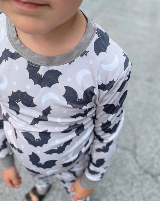 A young child wearing the Batty • 2 Piece Bamboo Set (Classic Fit), featuring black bats, white moons, and stars, stands outside on a paved surface. The image highlights the upper body with their face partially visible.