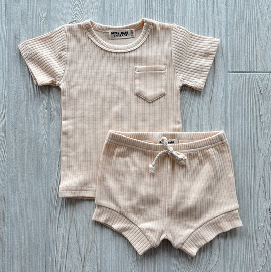 The Bone • Ribbed Shorty Set includes a beige ribbed organic cotton short-sleeve shirt with a chest pocket and matching drawstring shorts, both displayed flat on a light wood surface.