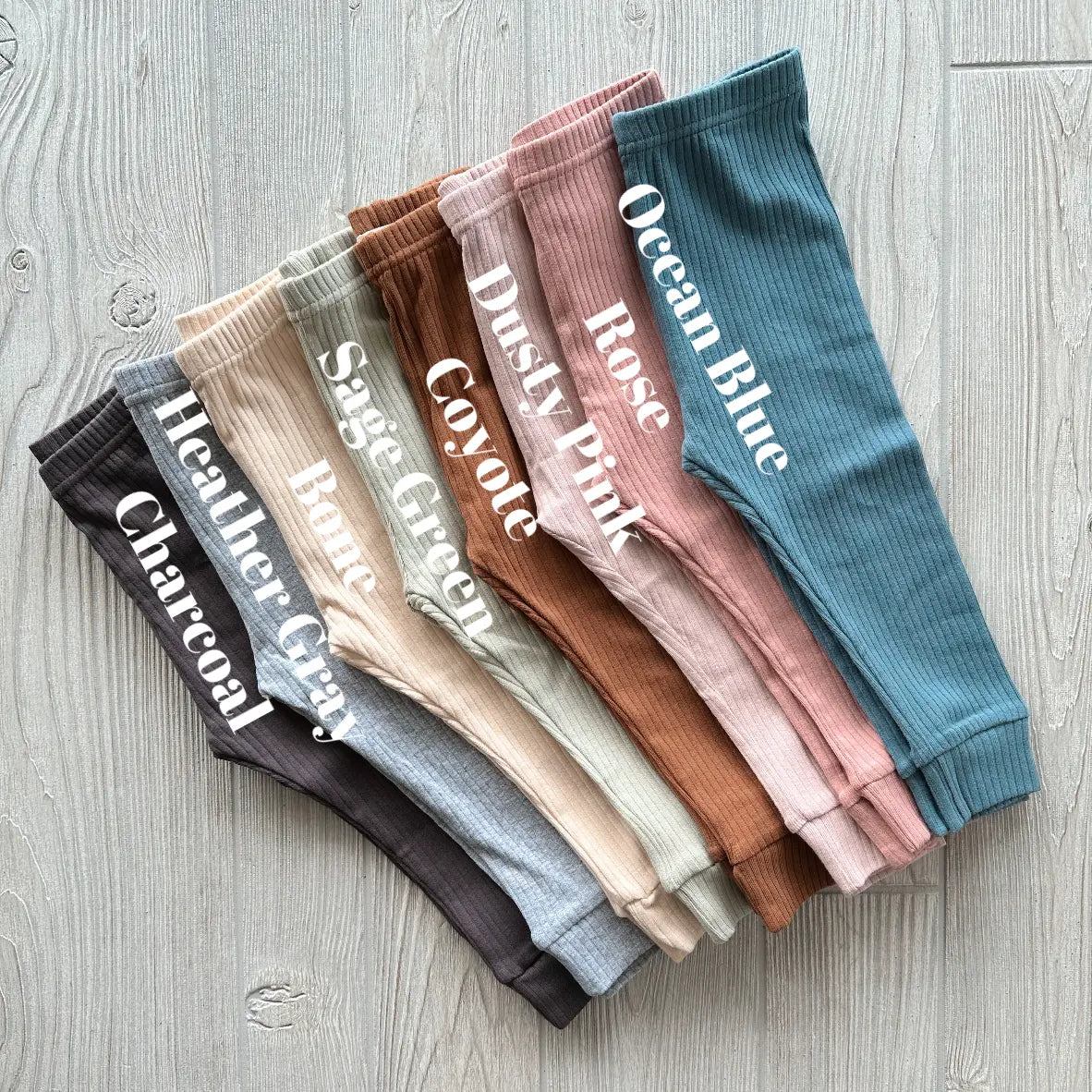 Eight pairs of Ribbed Organic Cotton Leggings—Charcoal, Heather Grey, Sage Green, Bare, Coyote, Dusty Pink, Rose, and Ocean Blue—are neatly arranged on a light wood surface as versatile base layers or stylish separates.