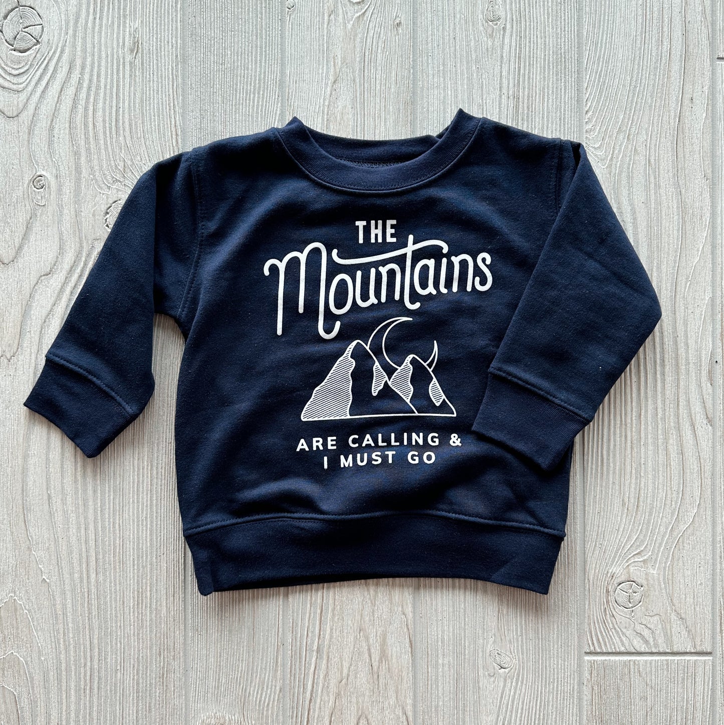 The Mountains are Calling • Navy Kids Pullover features white text and graphics with mountains and a crescent moon, laid flat on a light wooden surface; available in comfy unisex sizing.