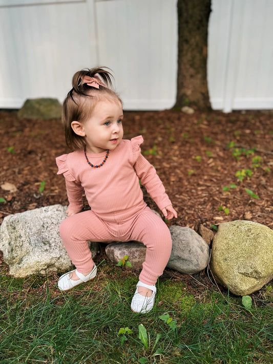 A toddler wears the Ribbed Ruffle Bodysuit in pink, paired with white shoes and a matching hair bow, sitting on outdoor rocks before a tree and white fence—a stylish organic cotton essential for cooler days.