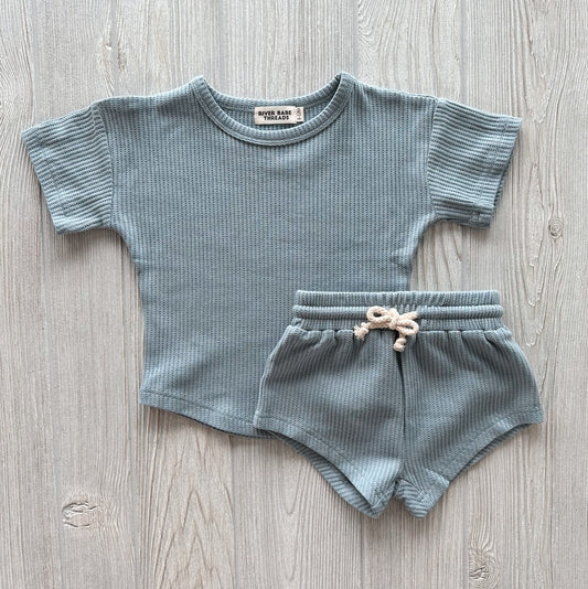 The Steel Blue • Waffle Shorty Set for babies and toddlers, featuring a subtle waffle texture, includes a short-sleeve shirt and drawstring shorts laid flat on a light wood surface. Fits true to size.