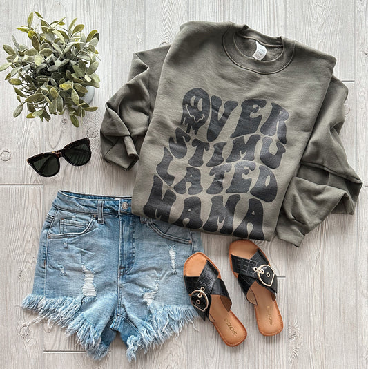 A flat lay of the Overstimulated Mama Olive Pullover paired with distressed denim shorts, black sunglasses, tan sandals, and a small potted plant on a light wood floor.