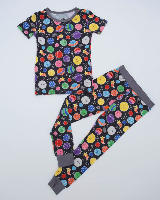 The Happy Planets • 2 Piece Bamboo Set (Classic Fit) features a space-themed kids’ pajama set with cheerful planets and stars, made from stretchy bamboo fabric, including a short-sleeve top and pants with gray cuffs and neckline.