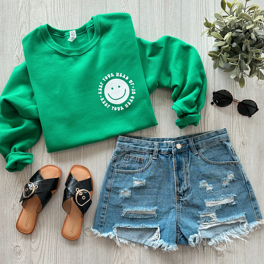 The Keep Your Head Up • Mental Health Pullover, a green unisex sweatshirt with a white smiling face and positive message, is styled with distressed denim shorts, black sandals, and a leafy plant on a light wooden background.