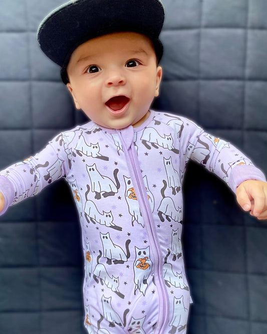 A smiling baby wearing the Ghost Cats • Bamboo Zippy (Classic Fit), a light purple onesie with white cartoon cat designs, stands against a dark quilted background and wears a black cap.