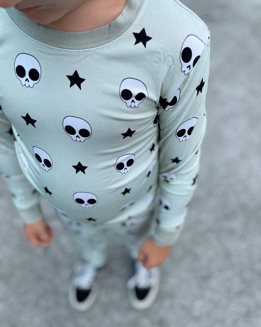 A child in the Rad Skelly • 2 Piece Bamboo Set (Classic Fit), a light green outfit with black skulls and stars made from bamboo viscose, stands on gray pavement with relaxed hands, wearing black-and-white sneakers.
