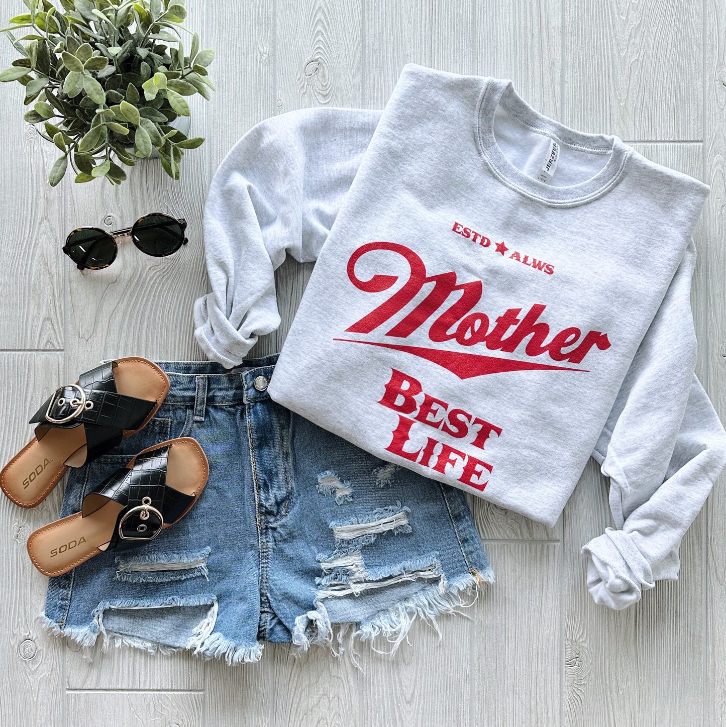 The Best Life • Ash Pullover—a vintage-inspired light gray unisex sweatshirt with red text—styled with ripped denim shorts, black slide sandals, sunglasses, and a small potted plant on a light wood floor.