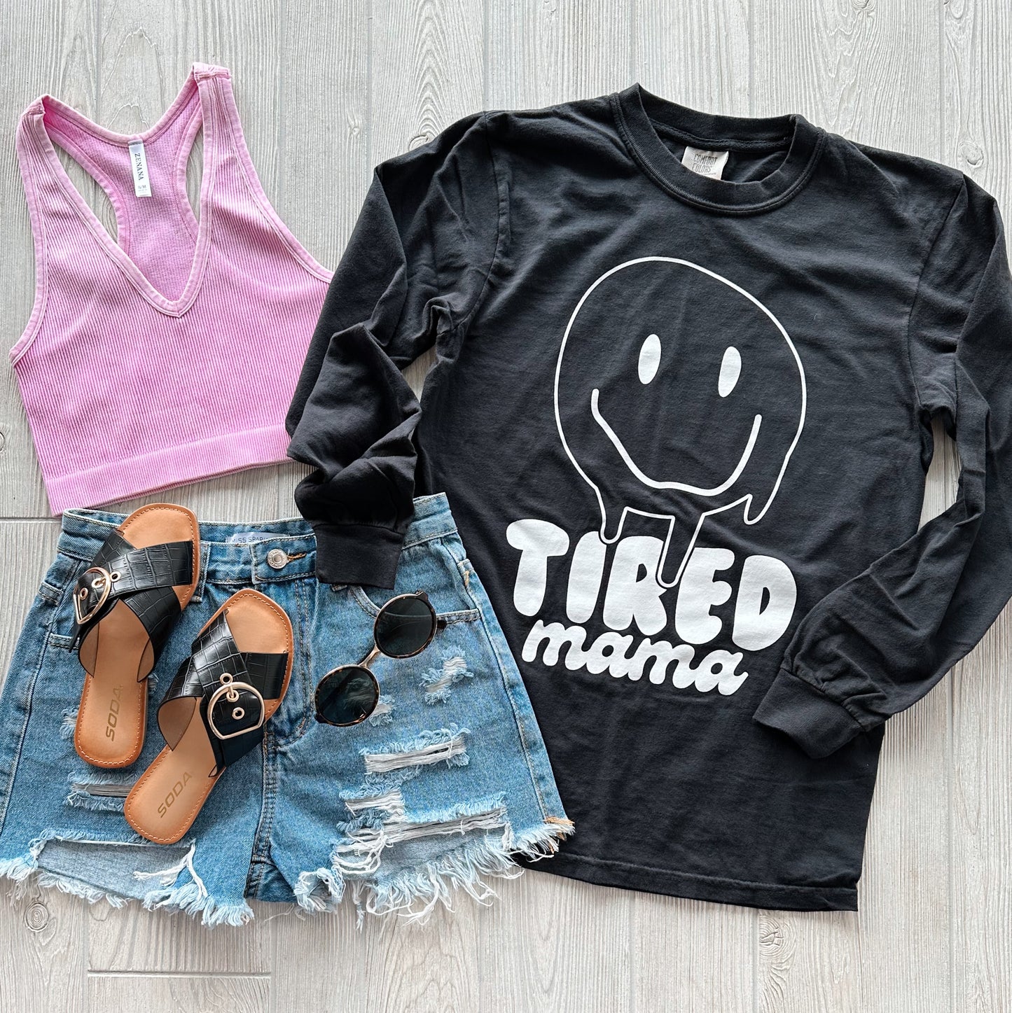 A flat lay of a pink ribbed crop top, Tired Mama • Vintage Black Long Sleeve tee with a smiling face graphic, distressed denim shorts, black sandals, and round sunglasses on a light wood floor.