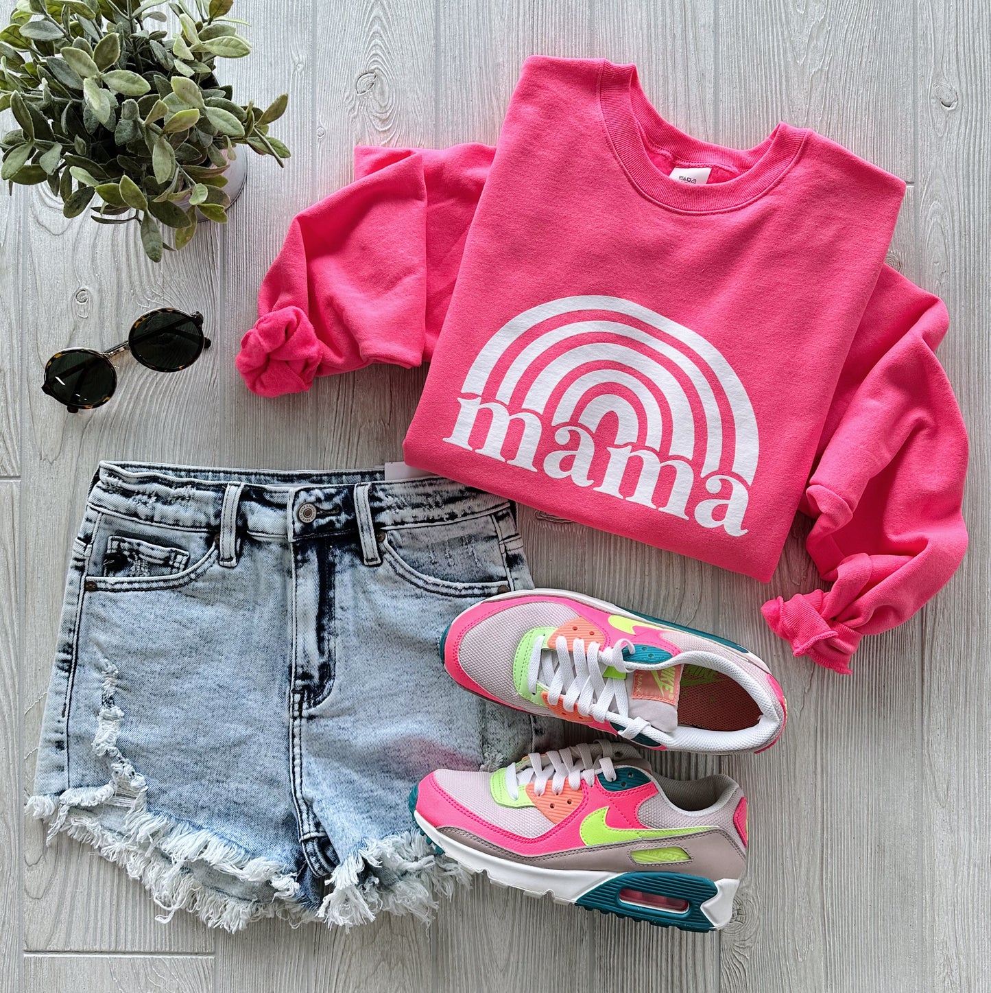 Flat lay of a casual outfit featuring the Mama Rainbow • Neon Pink Pullover, distressed denim shorts, colorful sneakers, black sunglasses, and a small green potted plant on a light wood floor.