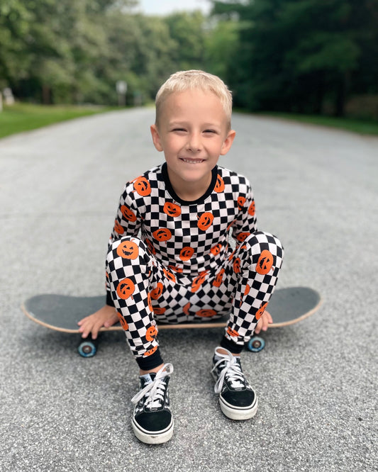 A smiling boy in the Checkered Pumpkins • 2 Piece Bamboo Set (Classic Fit) pajamas sits on a skateboard in the road, greenery behind him—super soft bamboo, perfect for eczema-prone skin.