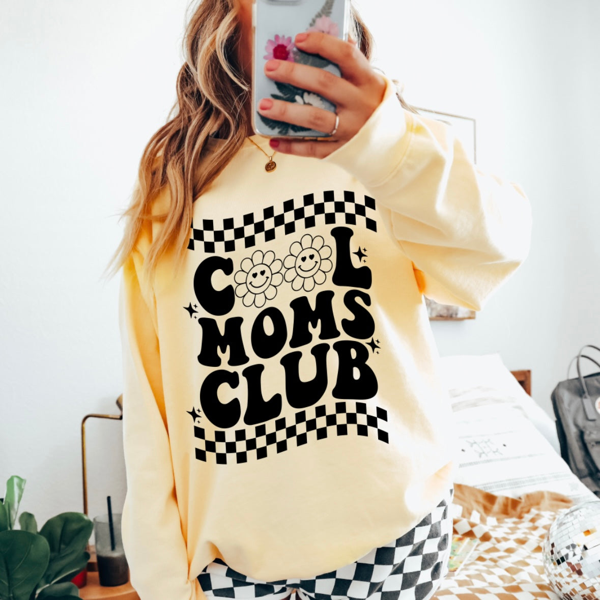A woman wears the Cool Moms Club • Butter Long Sleeve, featuring bold black lettering and flower designs, paired with black-and-white checkered pants as she takes a mirror selfie in a bright room.