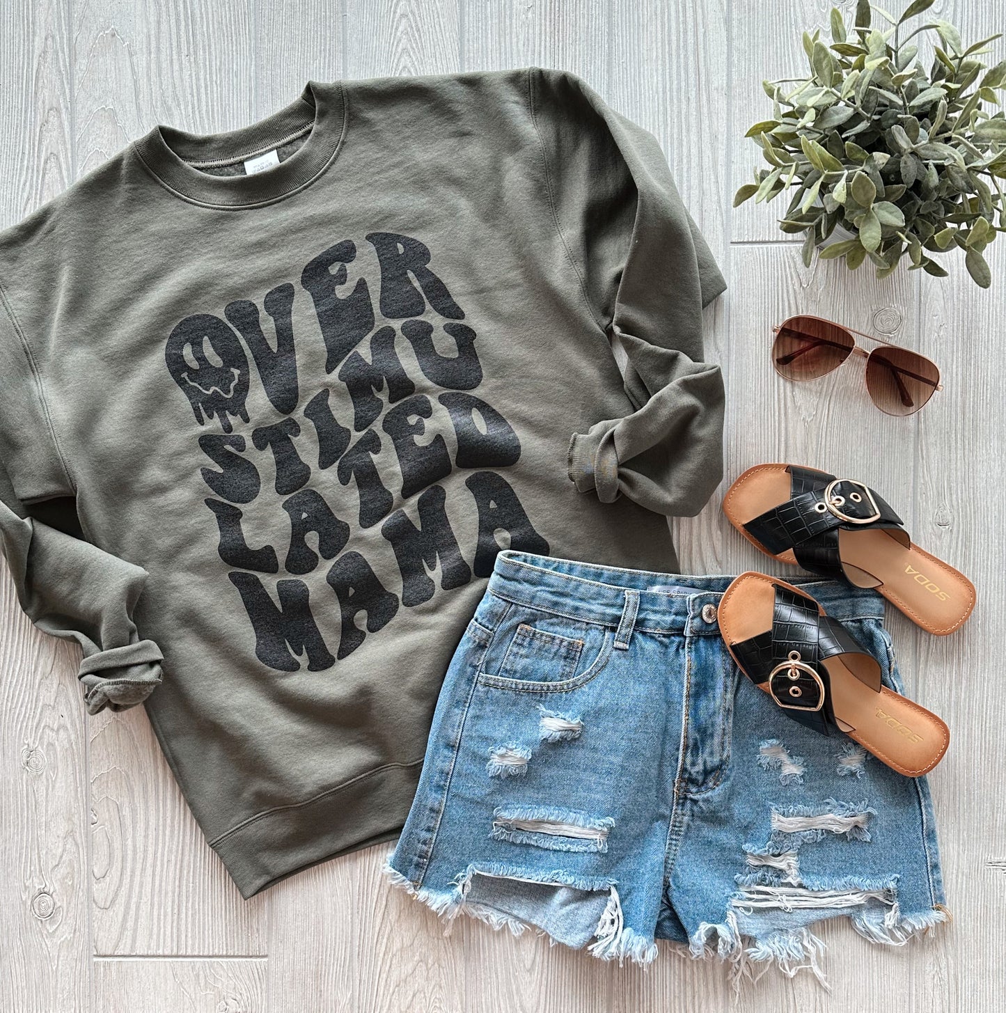 A styled flat lay with an olive Overstimulated Mama • Olive Pullover, distressed denim shorts, black sandals, brown sunglasses, and a potted plant on a light wood floor.