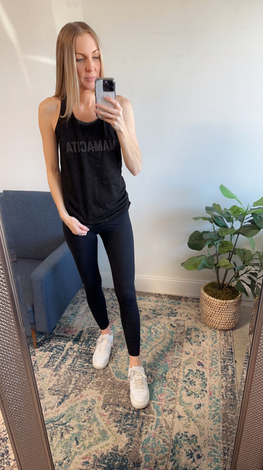 A woman takes a mirror selfie in a black MAMACITA • Unisex Tonal Tank, paired with black leggings and white sneakers. She stands on a patterned rug beside a blue chair and potted plant in a well-lit room.