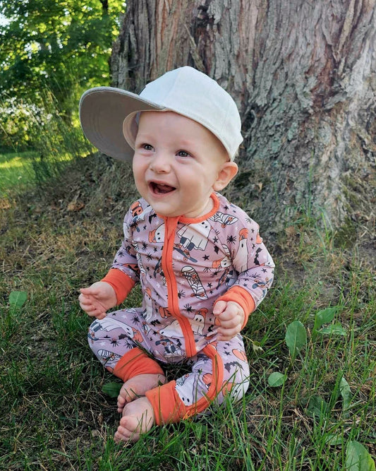 A smiling baby with sensitive skin wears the Skater Ghosts • Bamboo Zippy (Classic Fit), sitting barefoot on grass beneath a large tree as sunlight filters through green leaves in the background.