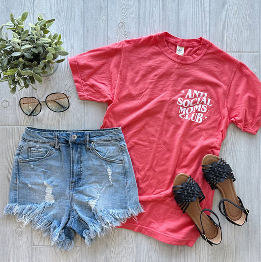 Flat lay of the Anti-Social Moms • Garment Dyed Tee in coral, featuring trendy text on ringspun cotton. Styled with distressed denim shorts, black sandals, sunglasses, and a small potted plant on a light wood background.