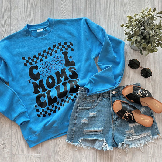 The Cool Moms Club • Blue Pullover features a unisex fit with bold black lettering, styled here with distressed denim shorts, black sandals, round sunglasses, and a potted plant on a light wood floor.