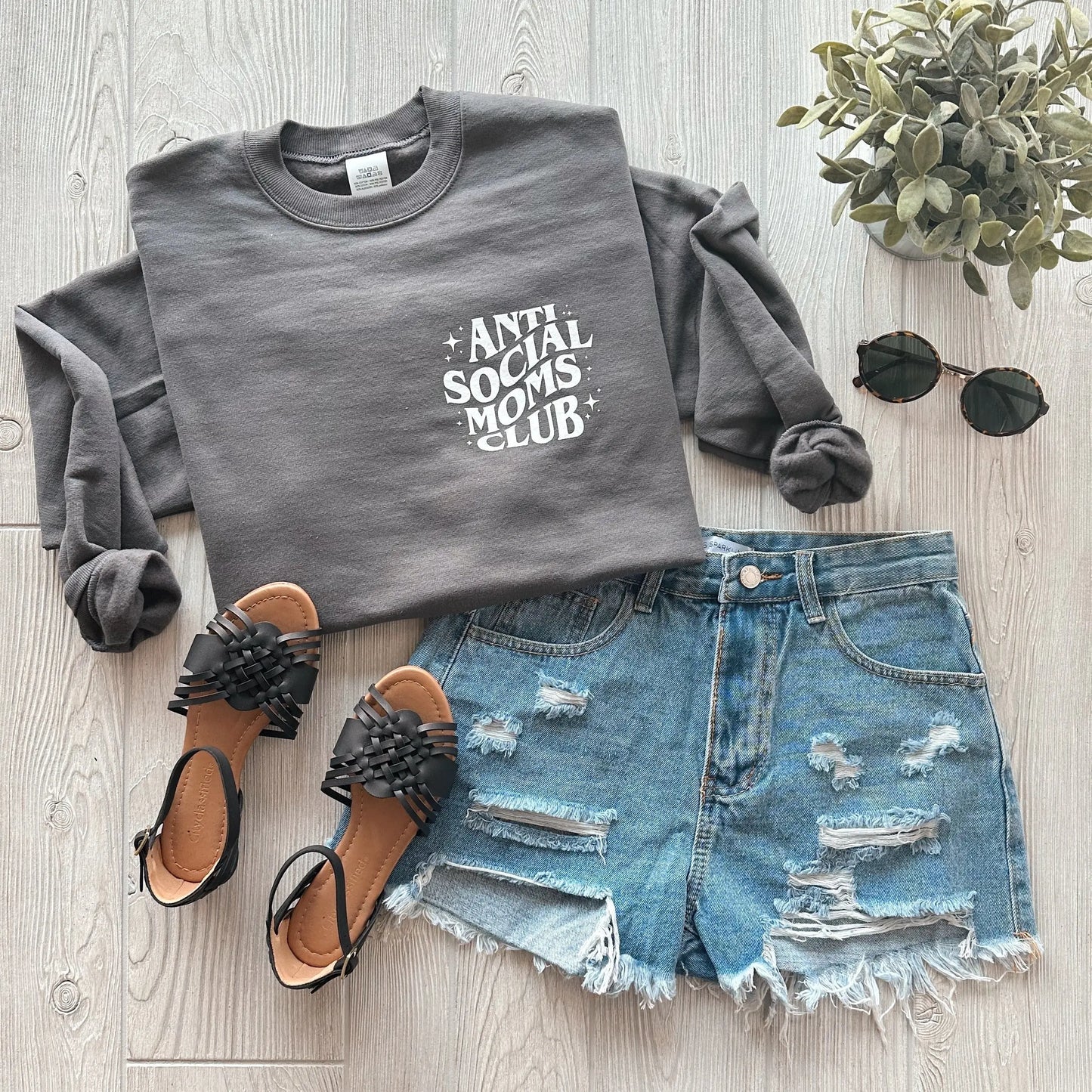 A flat lay of the Anti-Social Mom • Asphalt Pullover, a grey unisex sweatshirt made with soft recycled polyester and PrintPro XP fleece, styled with distressed denim shorts, black sandals, sunglasses, and a small green potted plant on light wood flooring.