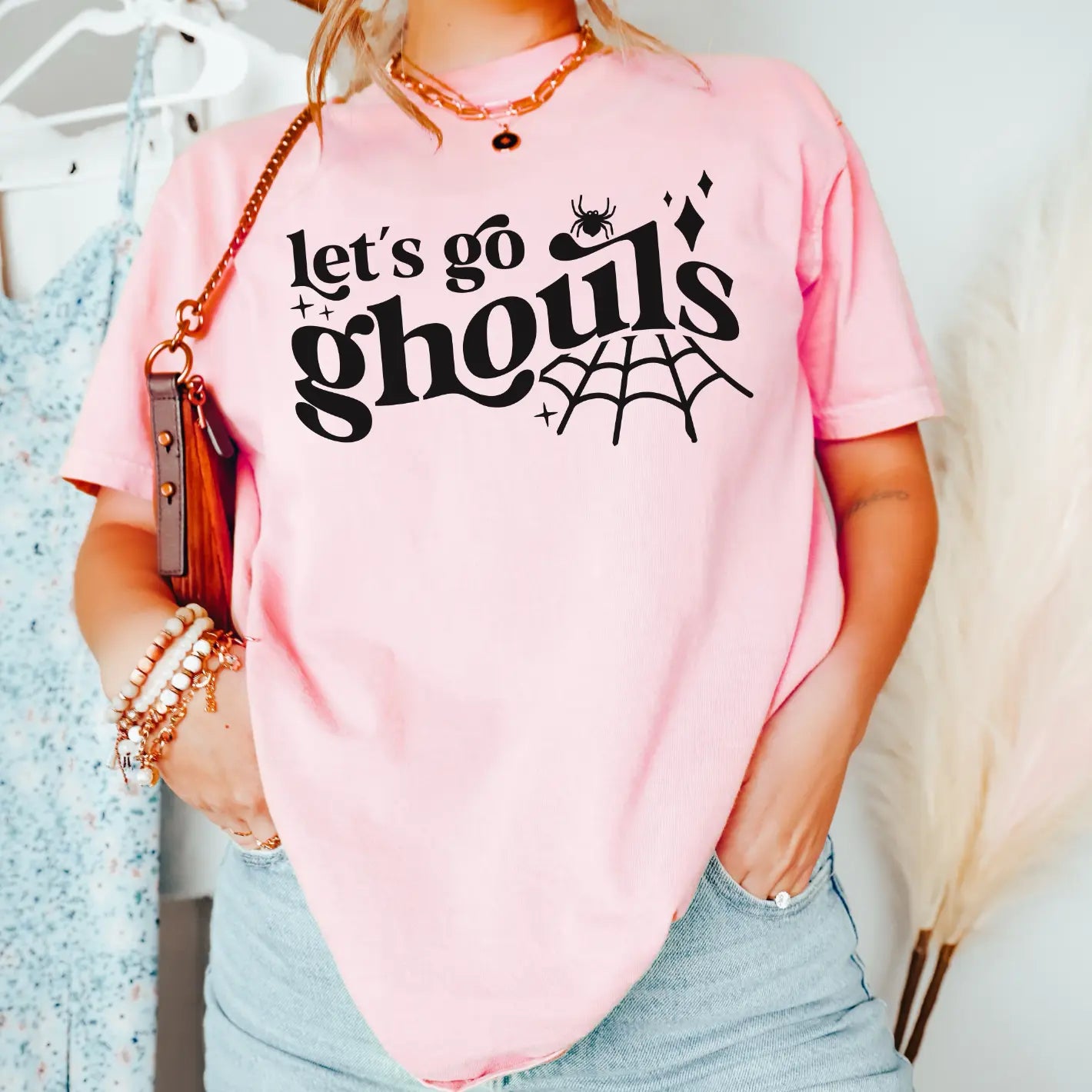 Person wears the Lets Go Ghouls pink tee, crafted from soft Airlume combed cotton with black text, spider, web, and sparkle graphics. Unisex sizing offers a retail fit. Styled with bracelets, purse, and one hand in pocket.