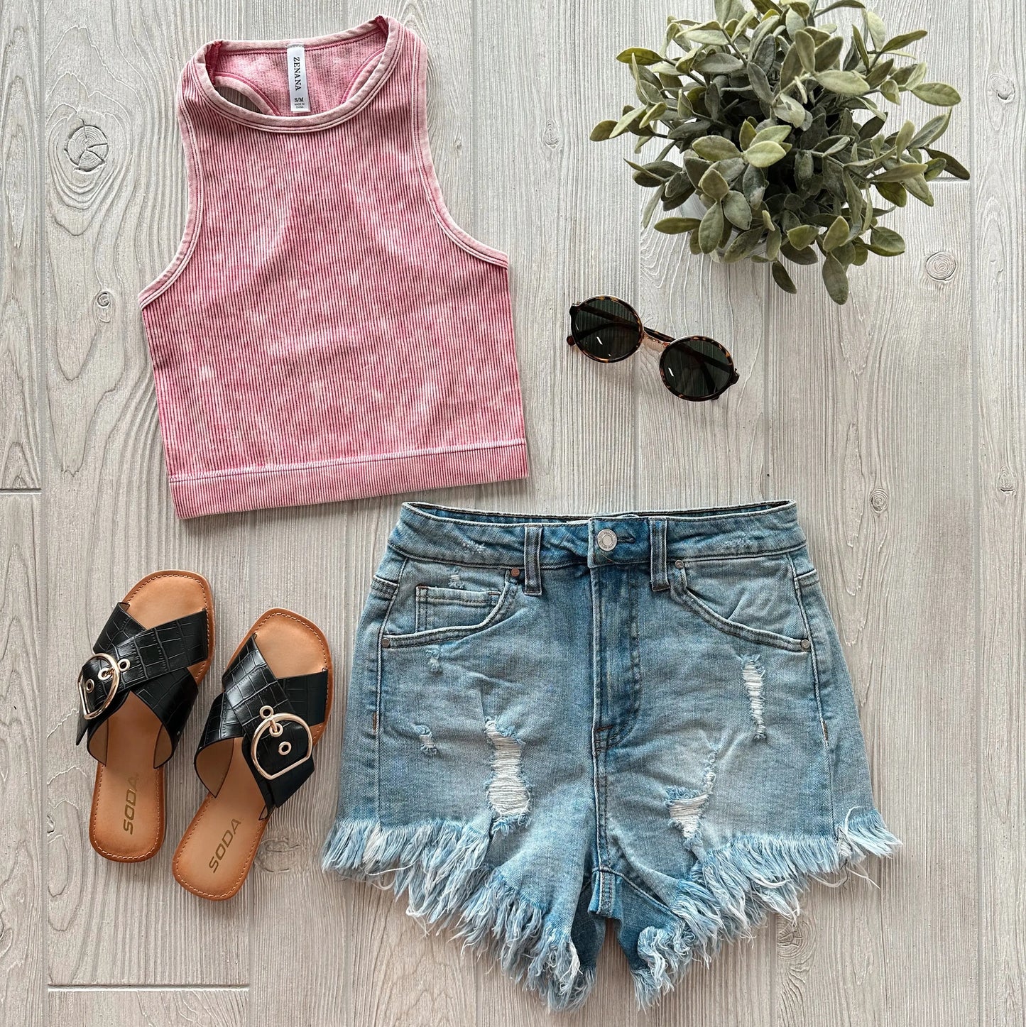 A flat lay of a summer outfit featuring the Sophia High-Neck Crop in Vintage Rose, distressed denim shorts, black slide sandals, round black sunglasses, and a small potted plant on a light wooden floor.
