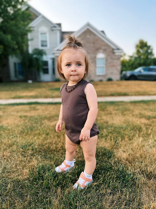 A toddler with light brown hair in a small ponytail stands on patchy grass, wearing an Organic Cotton Bubble Romper, a beaded necklace, and sandals. A brick house and car appear in the background.