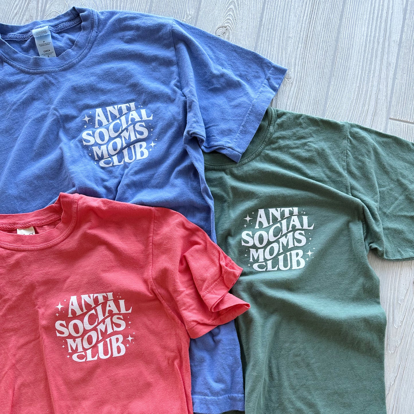 Three Anti-Social Moms • Garment Dyed Tees in blue, green, and red are displayed on a light wooden floor. Each shirt has Anti Social Moms Club in white stylized text on the front left and offers comfortable unisex sizing.