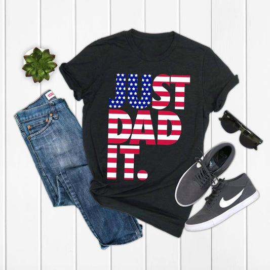 Flat lay of the JUST DAD IT • Flag Tee by Bella Canvas featuring bold American flag lettering, displayed with blue jeans, gray Nike sneakers, black sunglasses, and a small succulent.