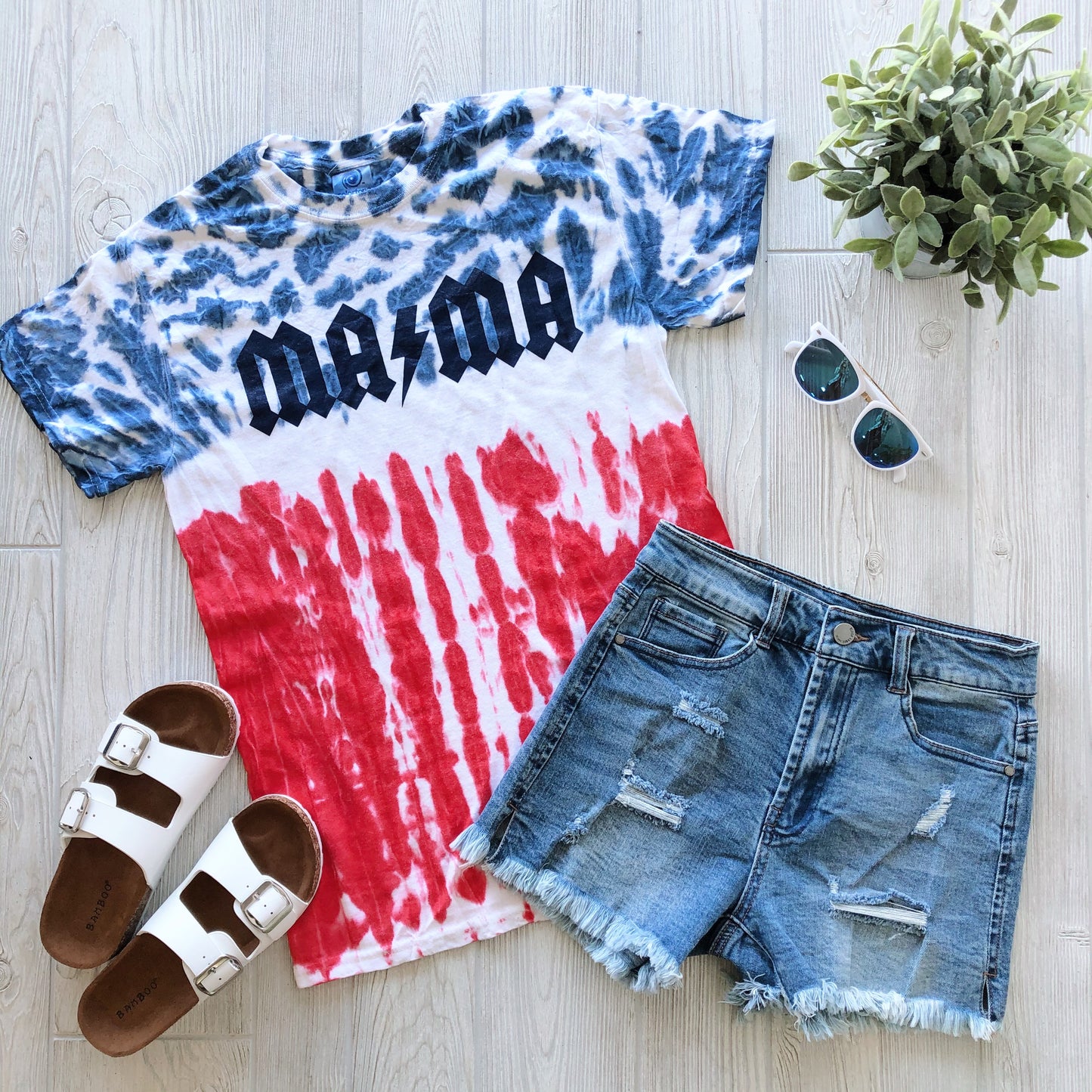 Flat lay of the MAMA AC/DC • Patriotic Tie-Dye t-shirt in blue, white, and red with distressed denim shorts, white sandals, sunglasses, and a small potted plant on a light wooden surface.